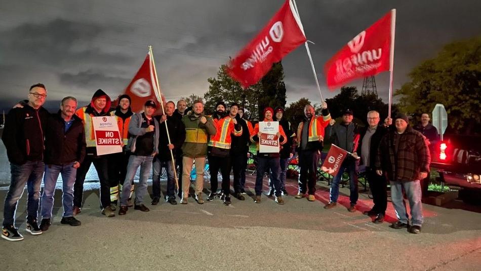 A large group of workers stand outside at midnight holding strike signs and flags